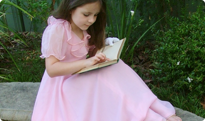 a photo of a young girl in a long pink dress, in a garden, reading a book